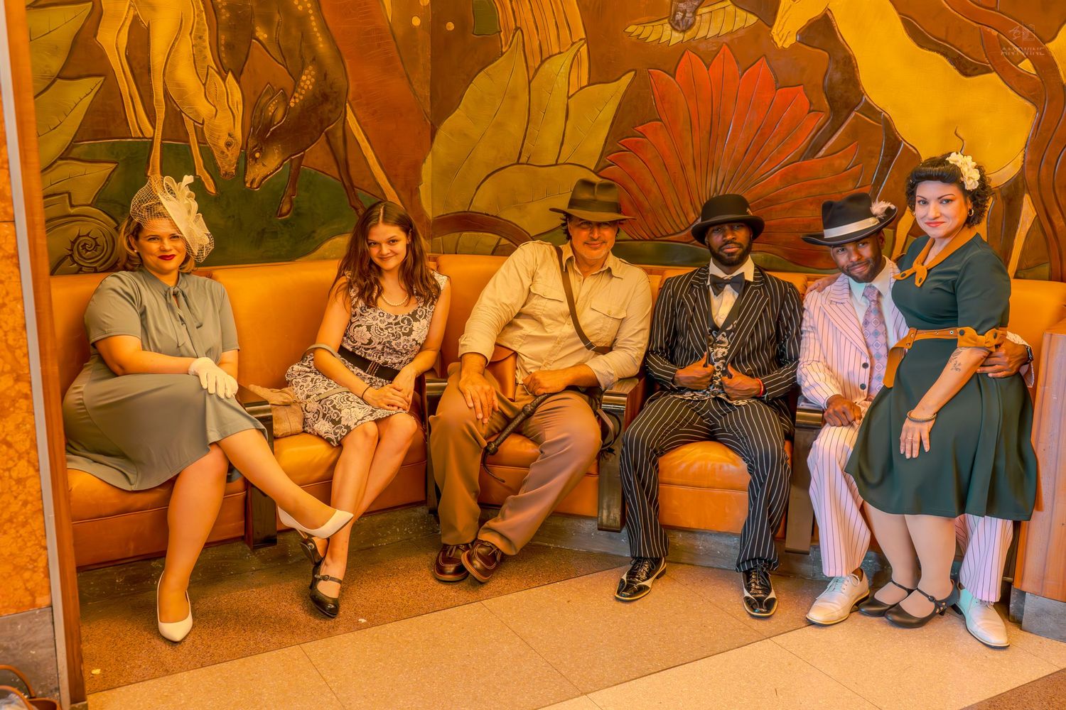 Six people in 1940s vintage attire sitting on an orange sofa in front of a colorful floral mural.