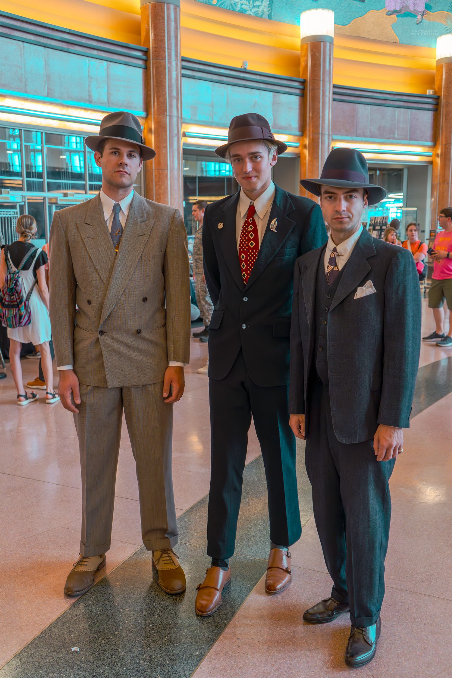 Three men in vintage 1940s suits and fedora hats posing together inside a grand building.