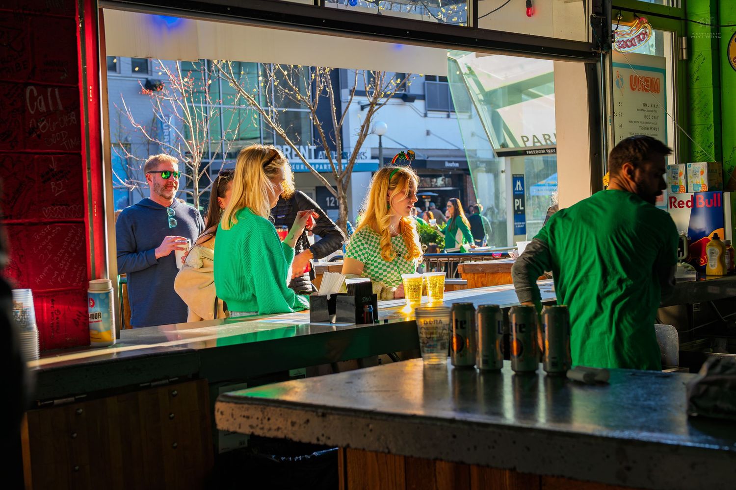 Bartender in green shirt serves drinks at a lively bar with patrons chatting near large windows at dusk.