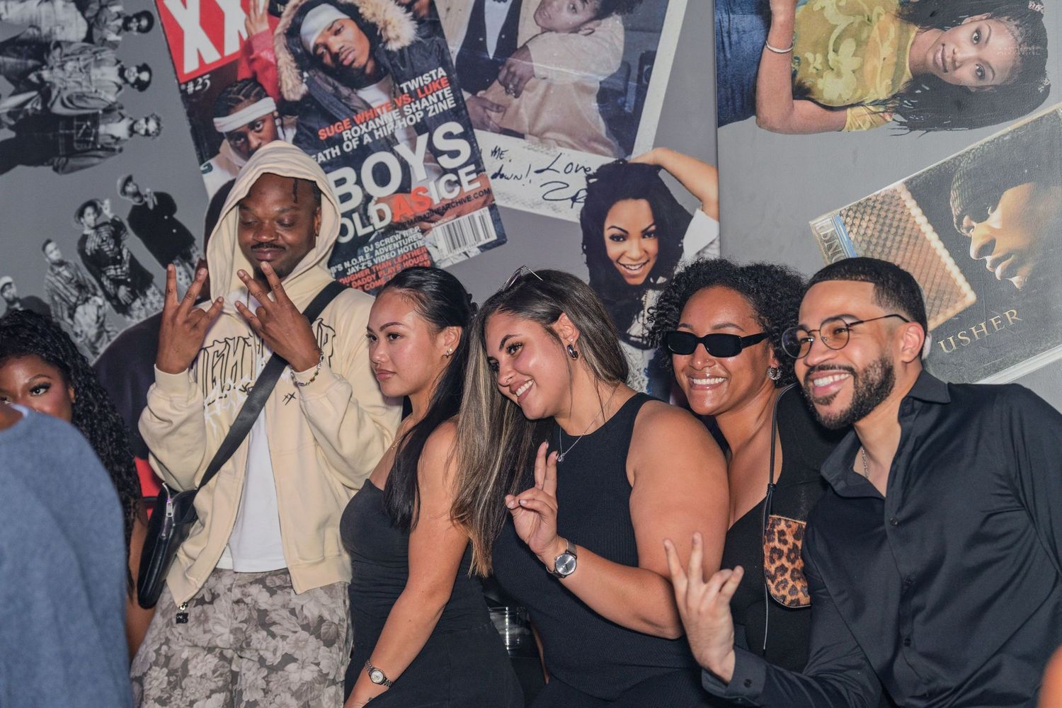 Group of friends posing together at a party with magazine covers displayed on the wall behind them.