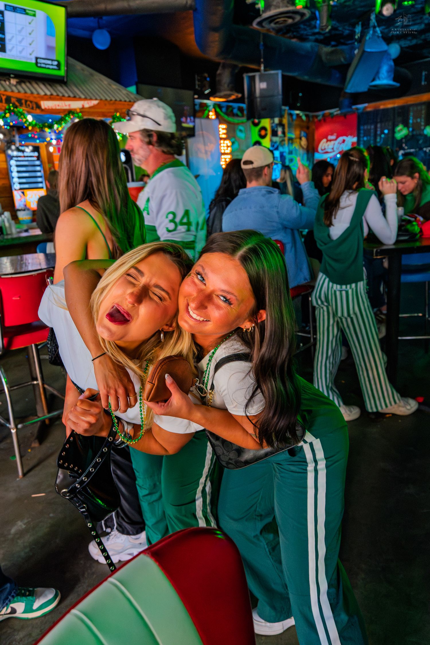 Two women in green outfits pose playfully at a lively bar during a St. Patrick's Day celebration.