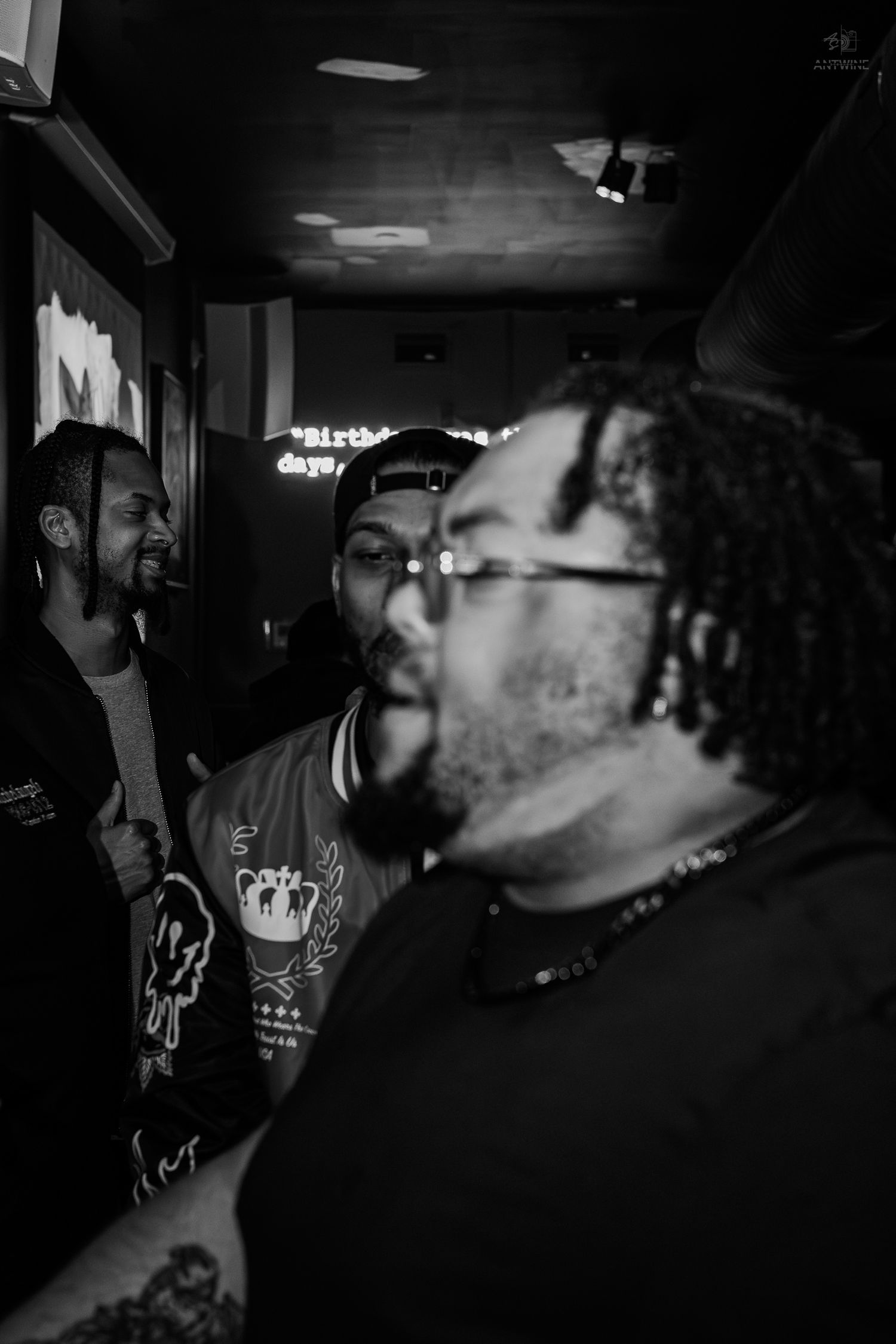 Three men conversing in a dark venue, one with dreadlocks and glasses wearing a graphic tee and beaded necklace.