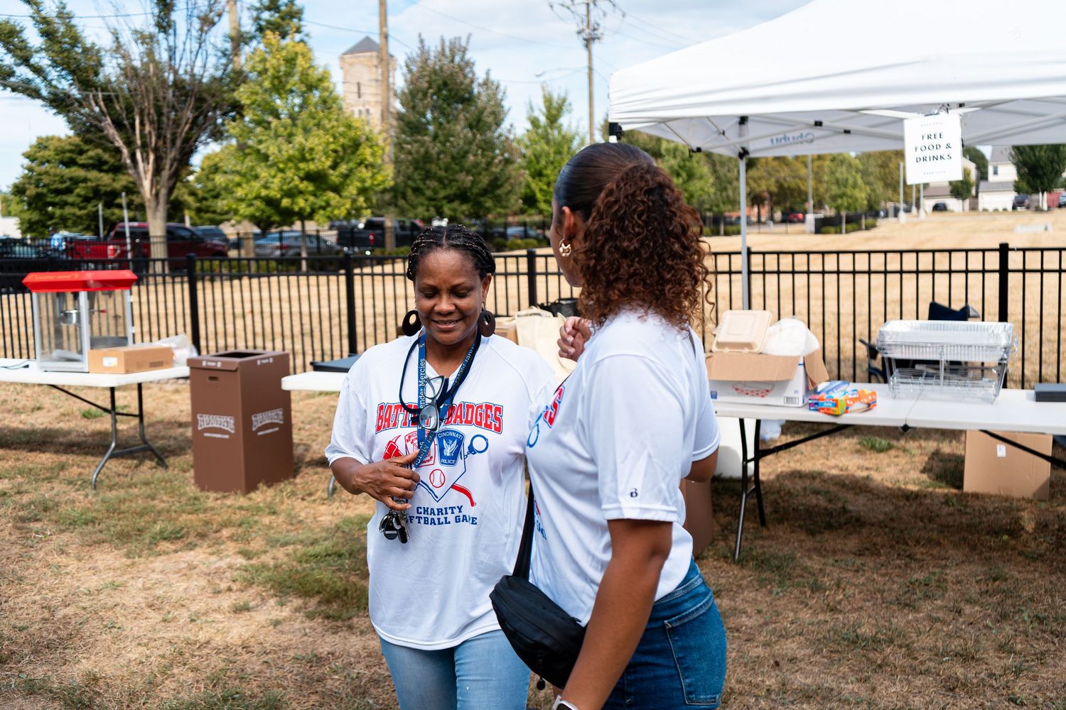 Two women in matching white t-shirts talking at an outdoor community event.