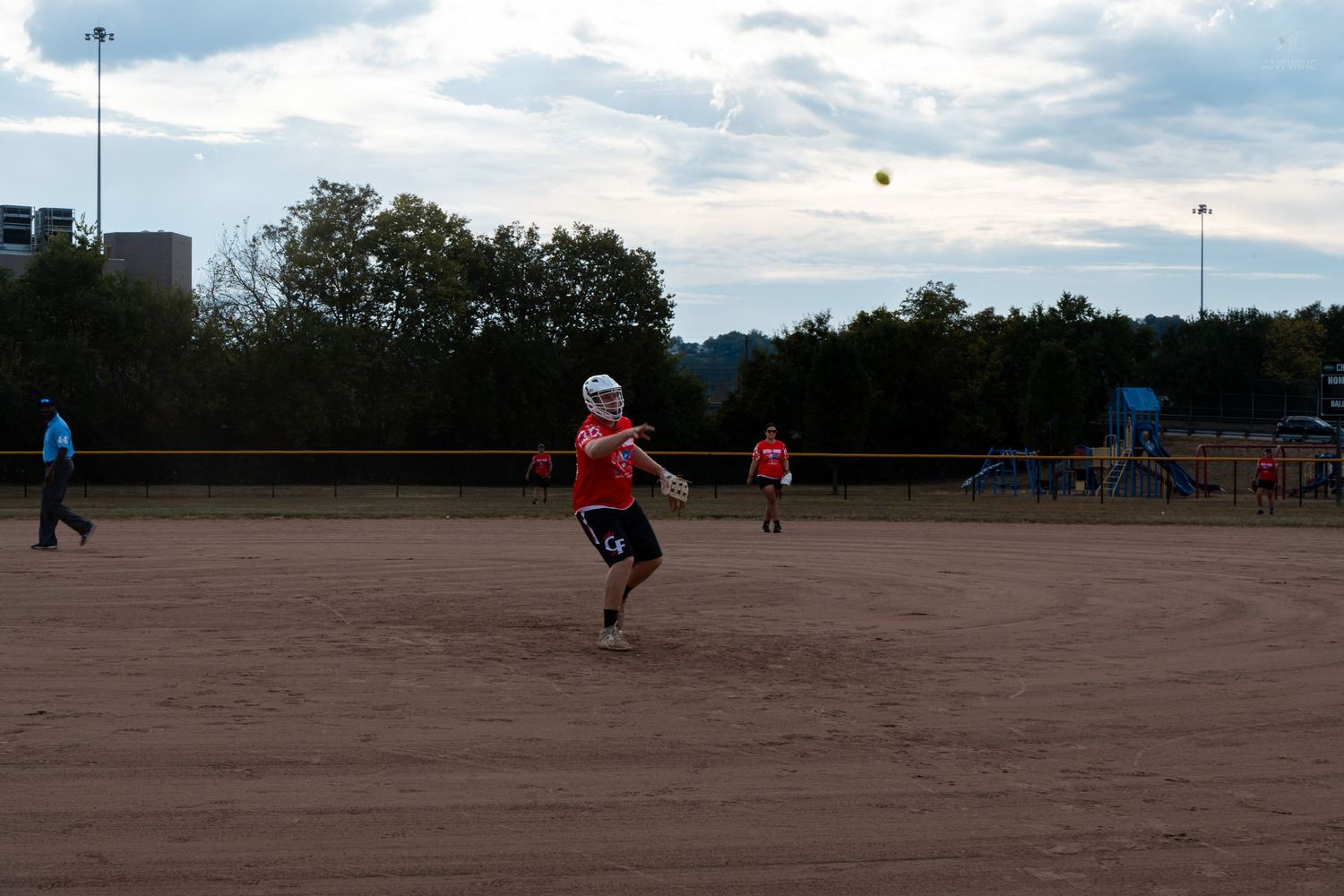 A softball player in a red jersey throws the ball across a dirt field during an evening game.