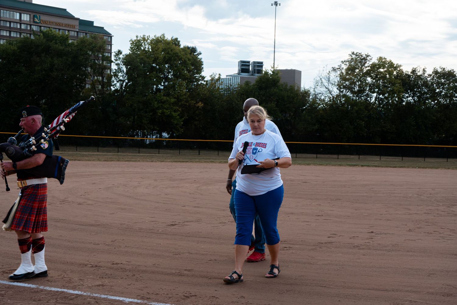 A woman in a white shirt walks on a dirt softball field while a bagpiper in a red tartan kilt plays nearby.