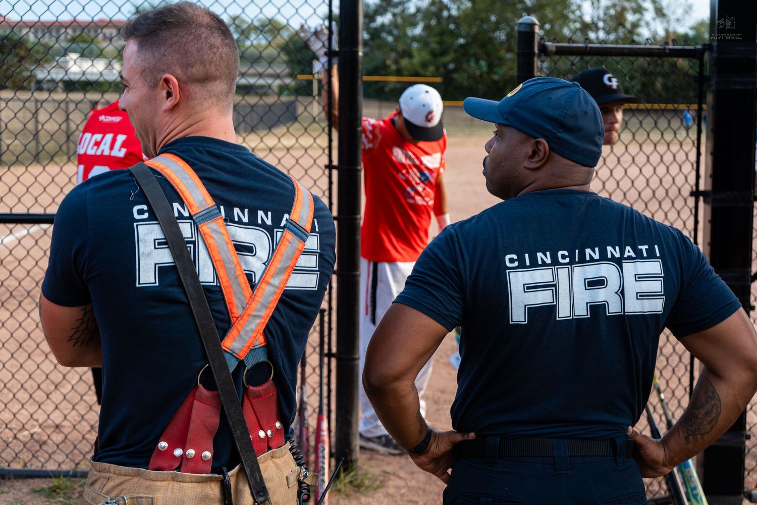 Two Cincinnati Fire firefighters in uniform black shirts watch a baseball game from behind a chain-link fence.