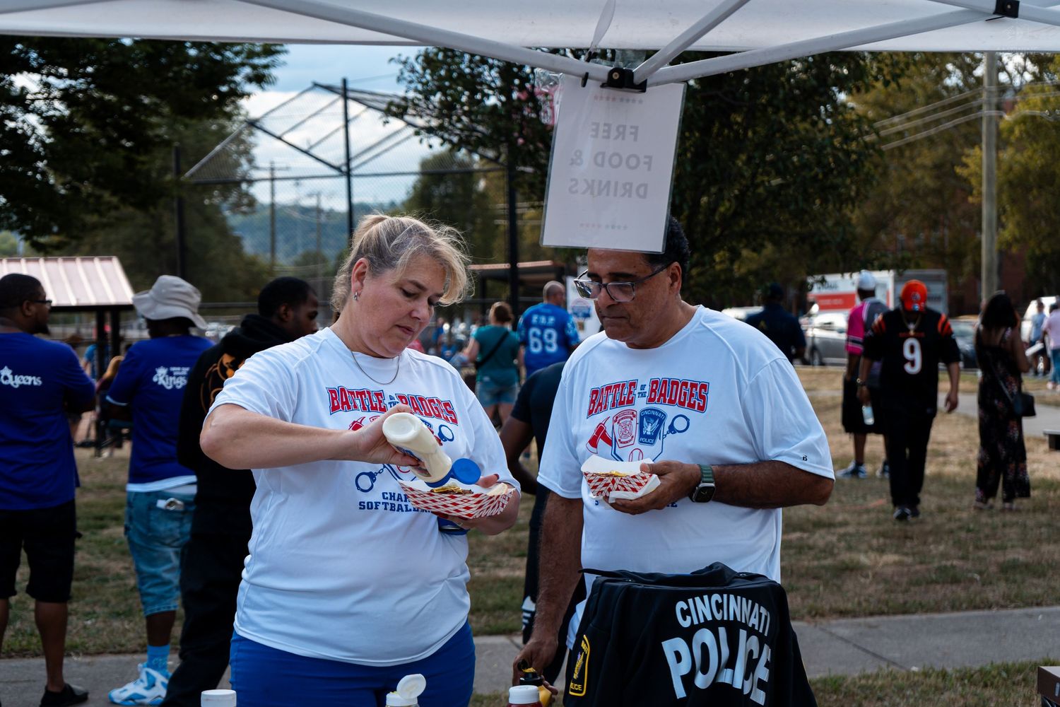 Two people in Battle of Badges shirts share food at an outdoor charity softball event tent.