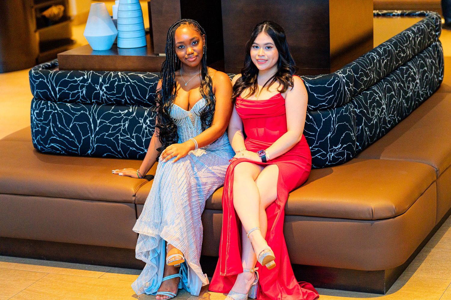Two young women in formal dresses posing together on a stylish sofa.