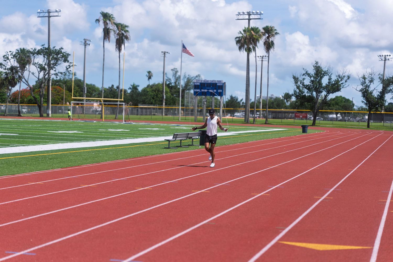 Athlete running on a red outdoor track and field stadium with palm trees and American flag.