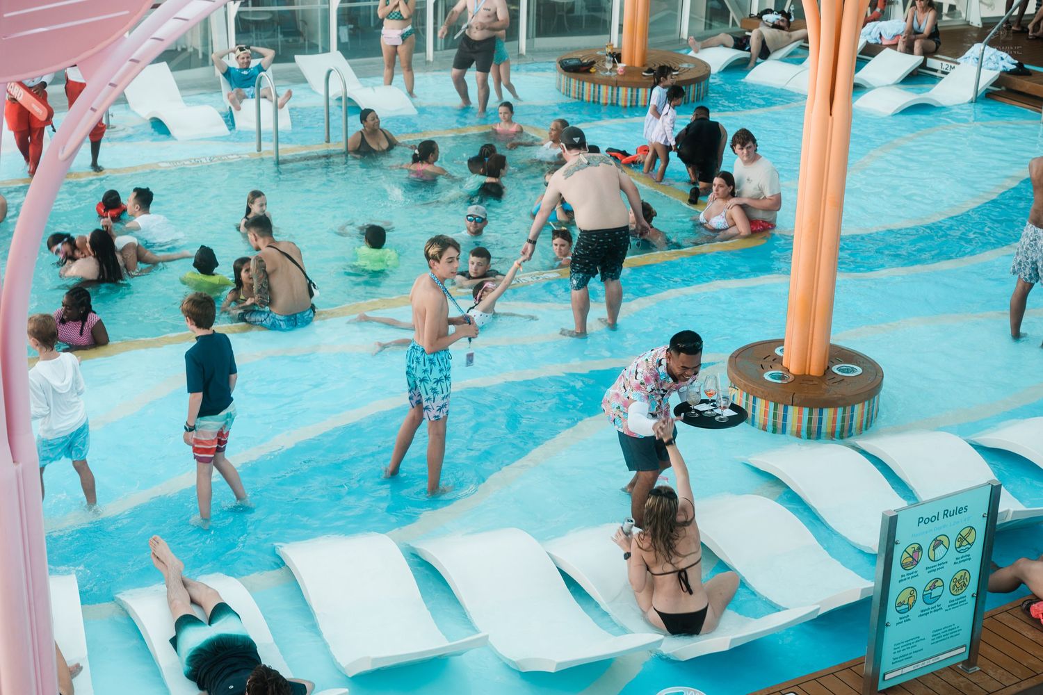 Overhead view of cruise ship guests swimming and relaxing in a bright blue pool area.