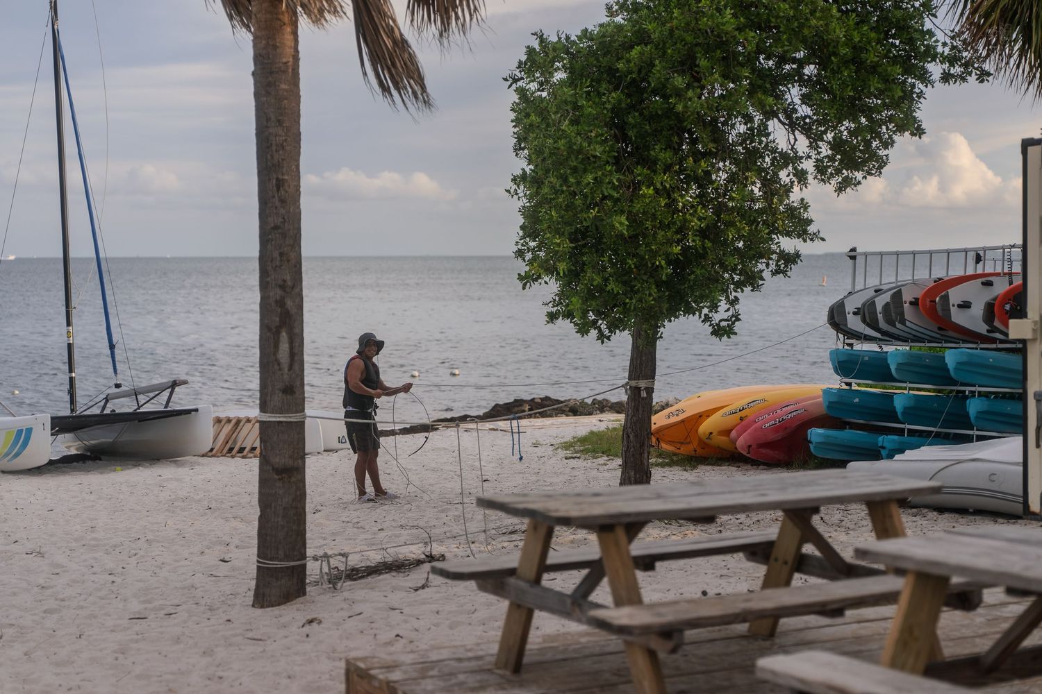 Beach rental area with kayaks, sailboat, and person preparing equipment at Key Biscayne shoreline.