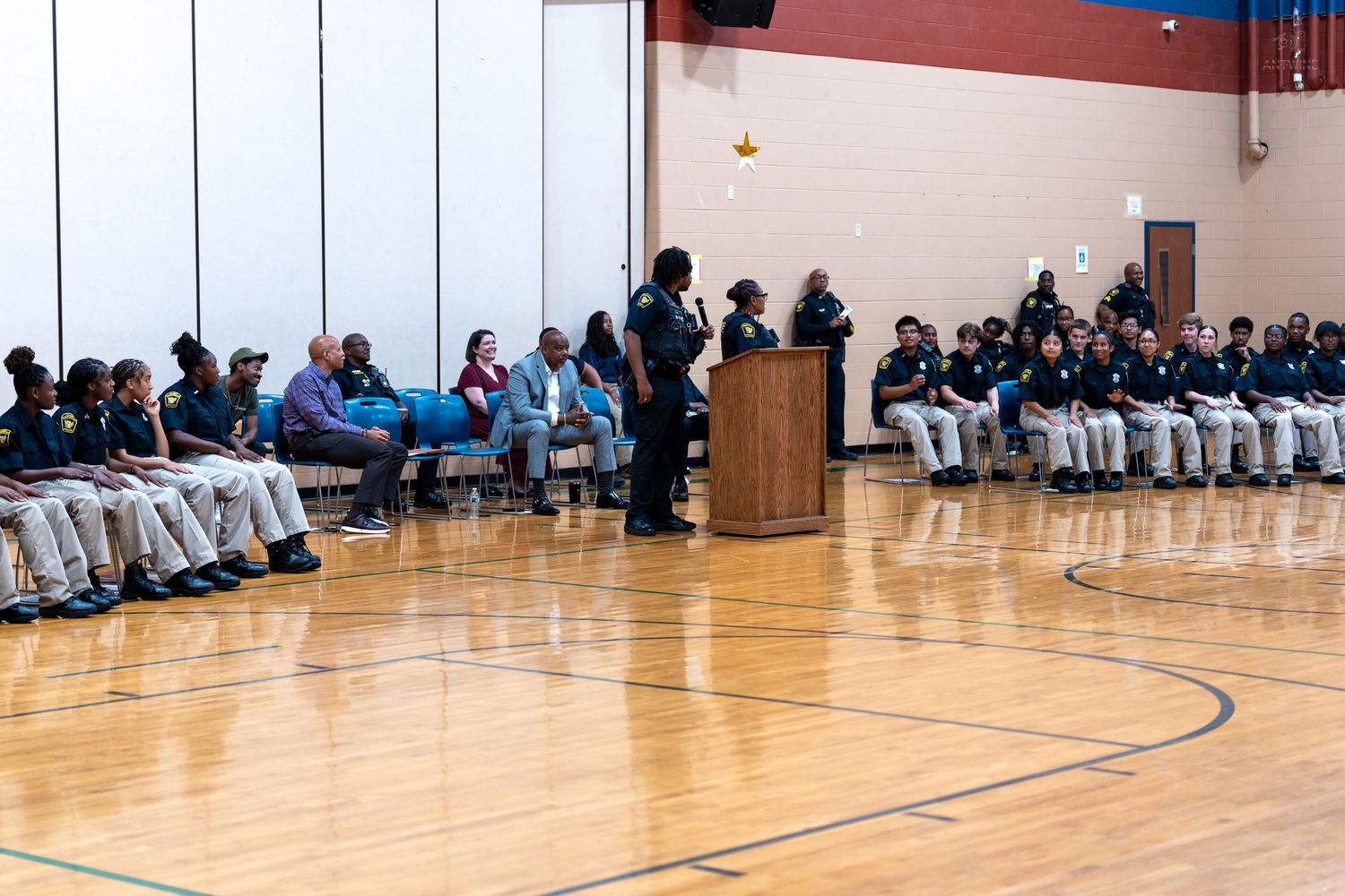 Speaker addresses seated students and staff in a school gymnasium during an assembly or awards ceremony.