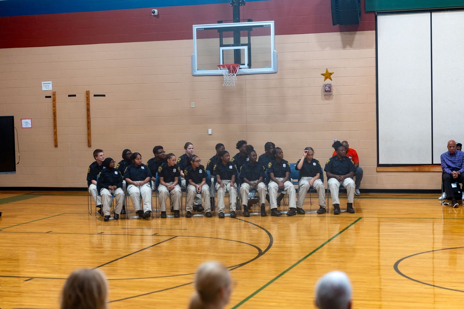 Group of youth in matching uniforms seated in a row on chairs inside a school gymnasium.