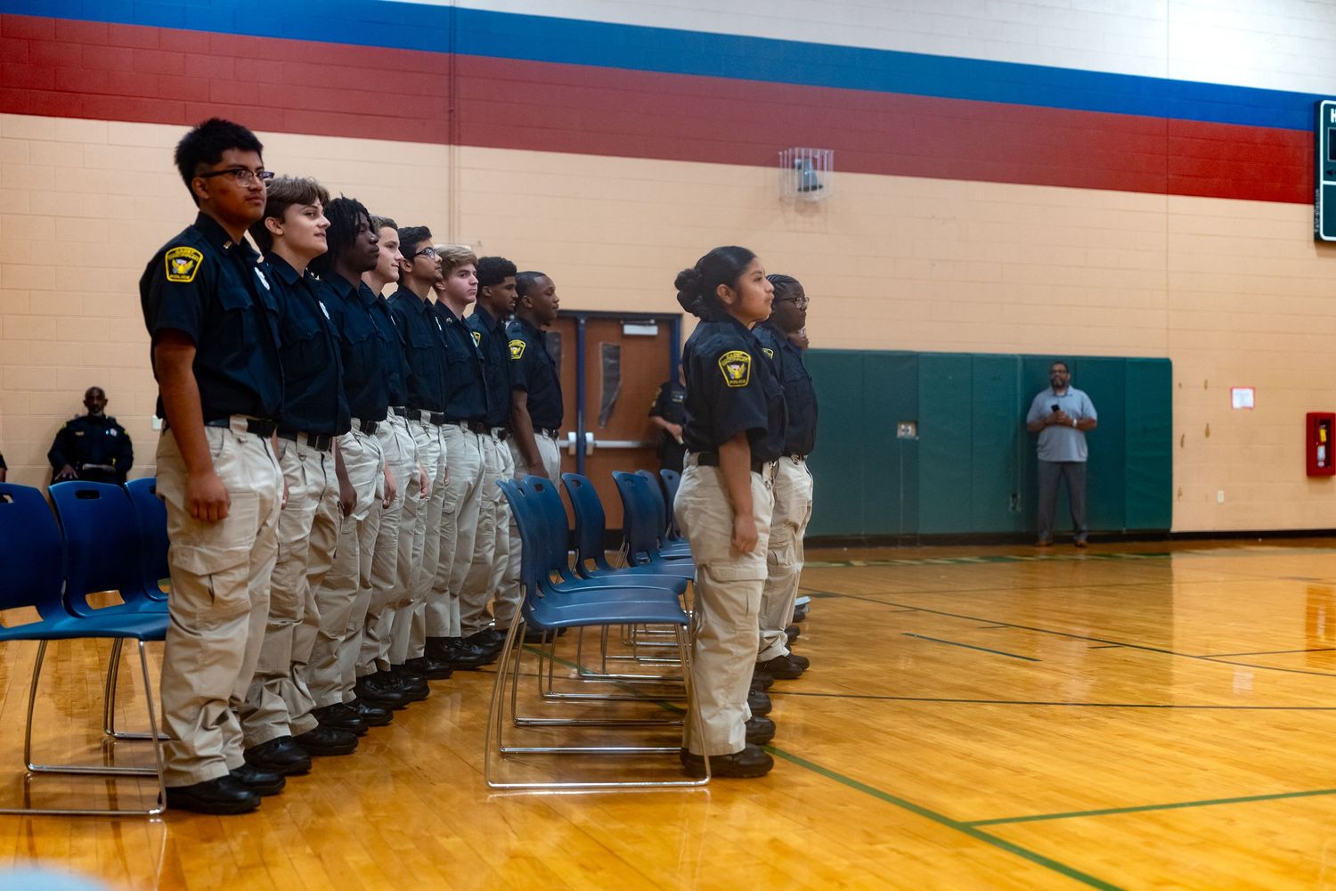 Young cadets in uniforms stand at attention in a school gymnasium during a formal ceremony or drill.