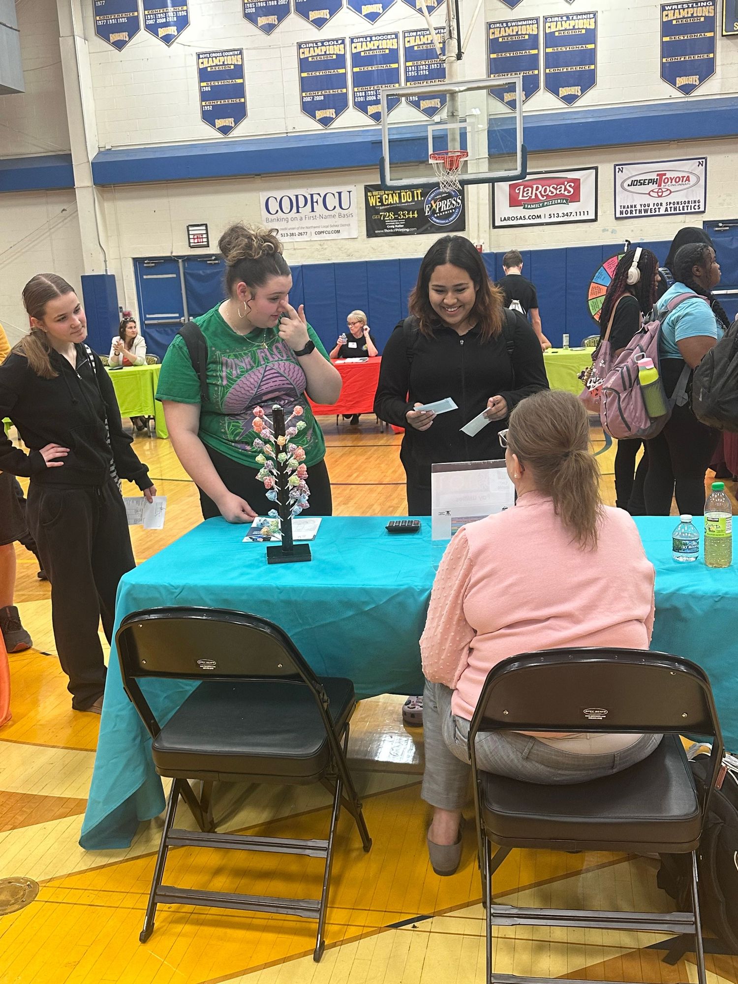 Students interact at a career fair booth in a school gymnasium with teal tablecloth and informational displays.