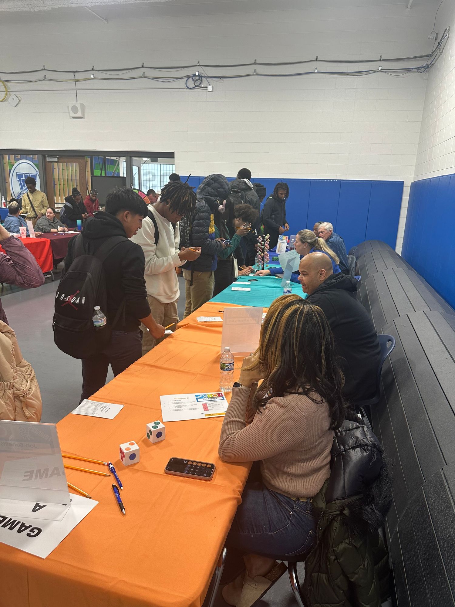 Students and attendees gather at orange tables during an indoor school career fair event in a gymnasium.