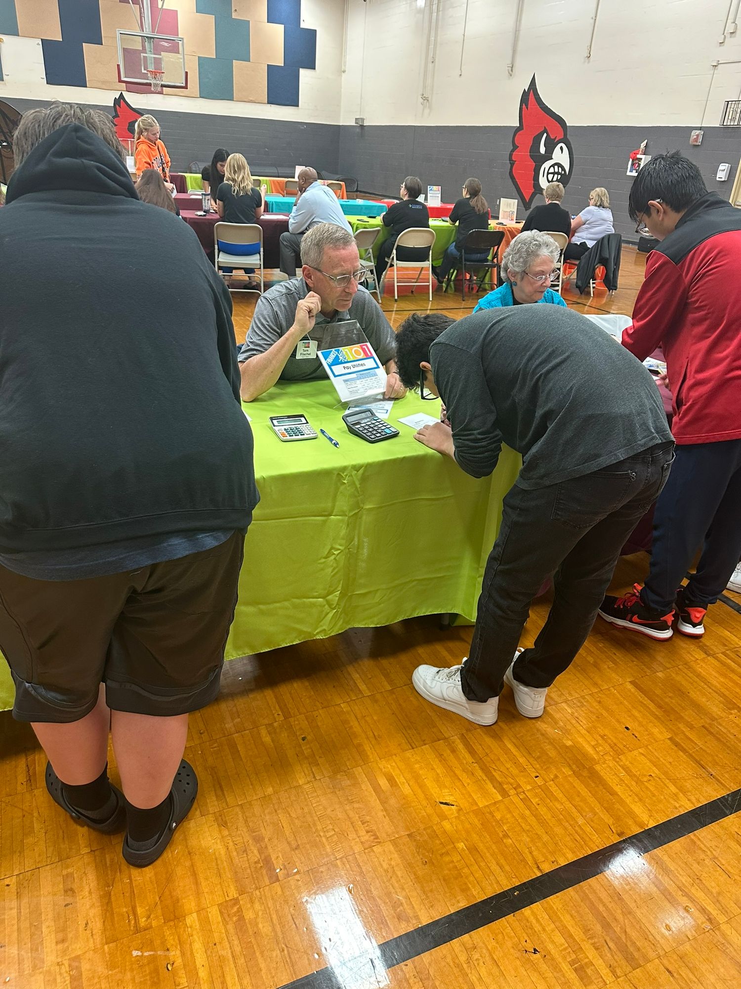 People gathered at green tables in a school gymnasium for a community event or job fair with Cardinal mascot on wall.