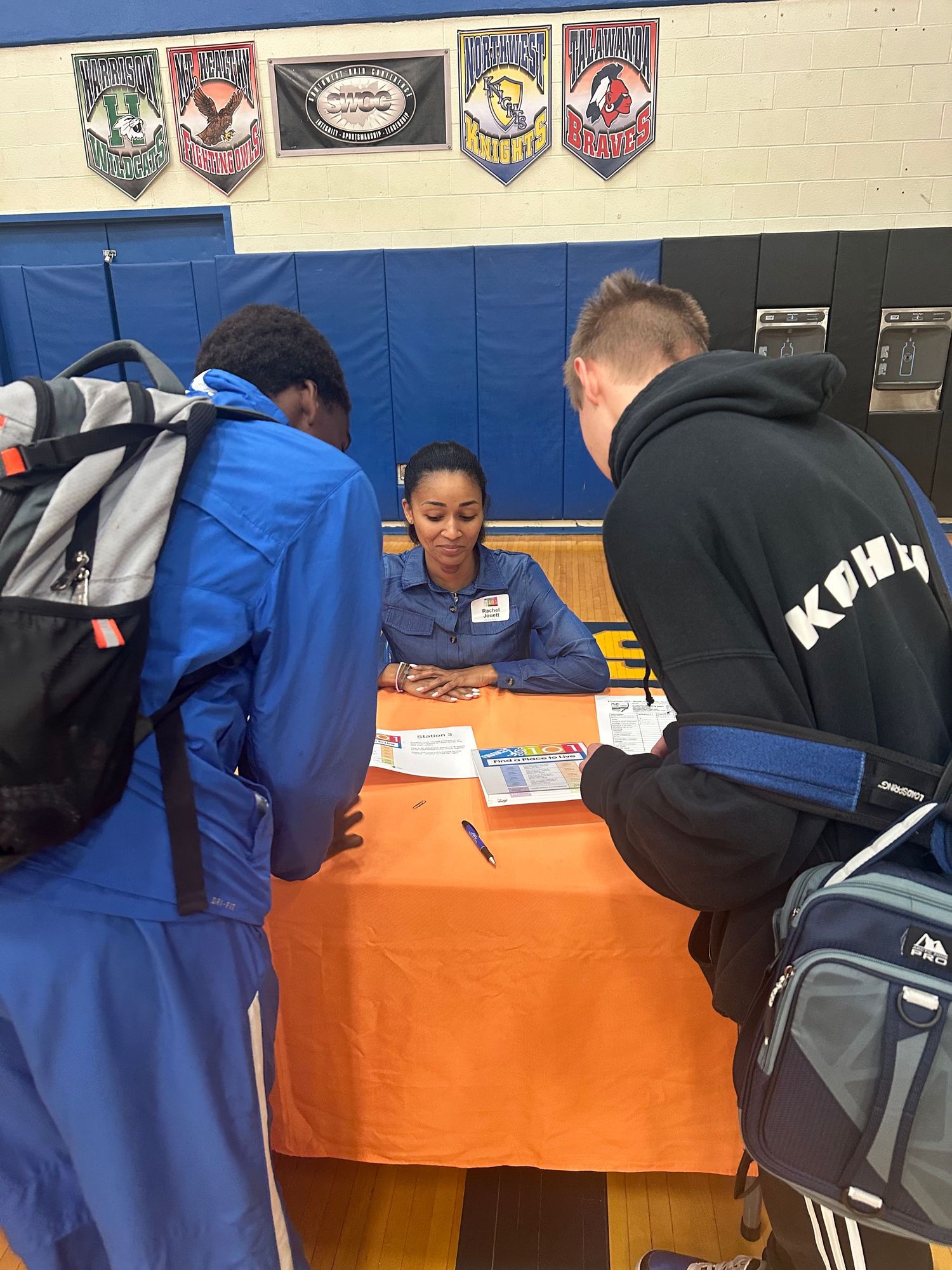 A woman sits at an orange table in a gym, helping two students with paperwork at a school career fair.
