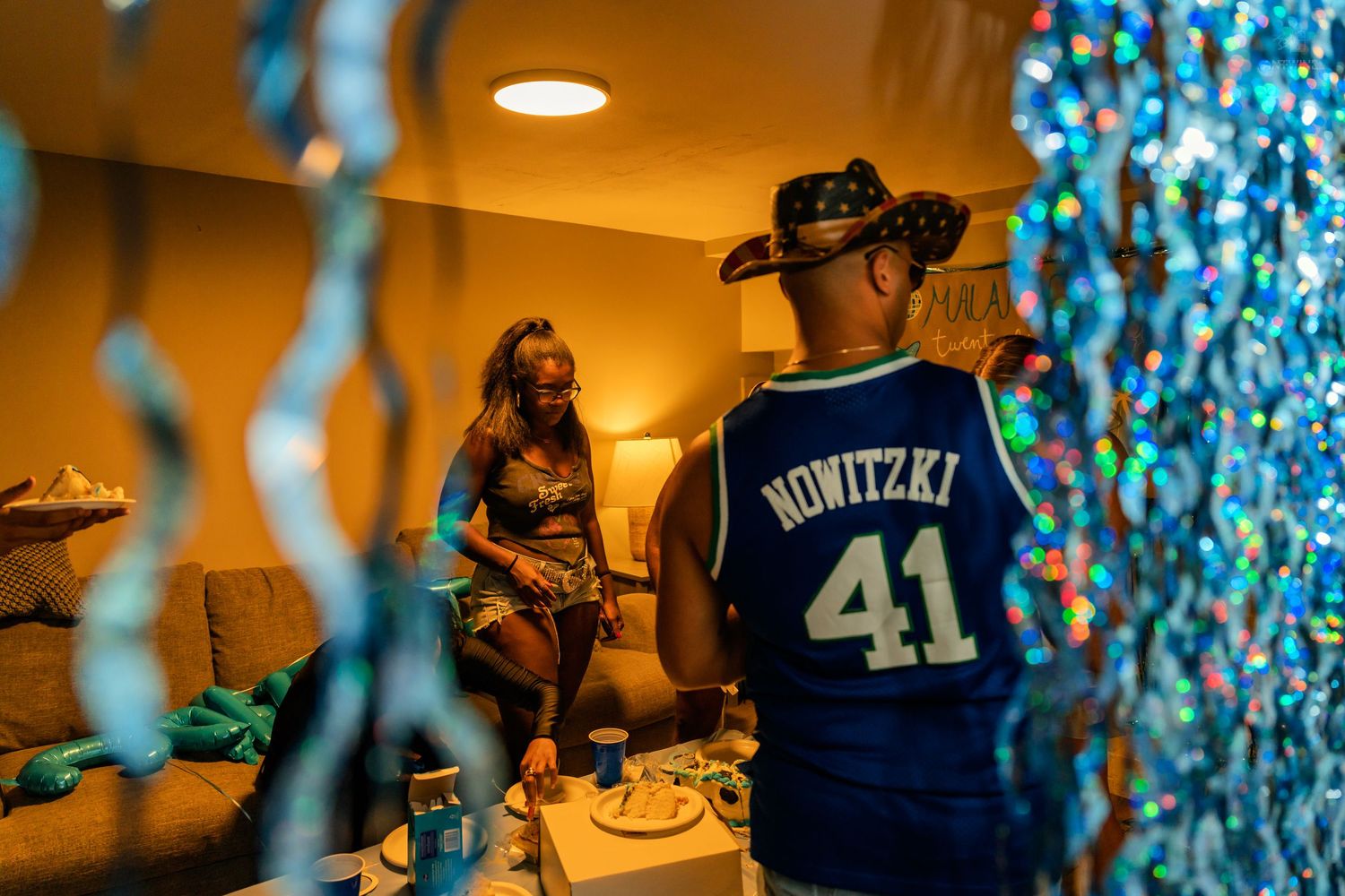 Man in Nowitzki 41 jersey chatting with a woman on a couch at a blue-themed birthday party.