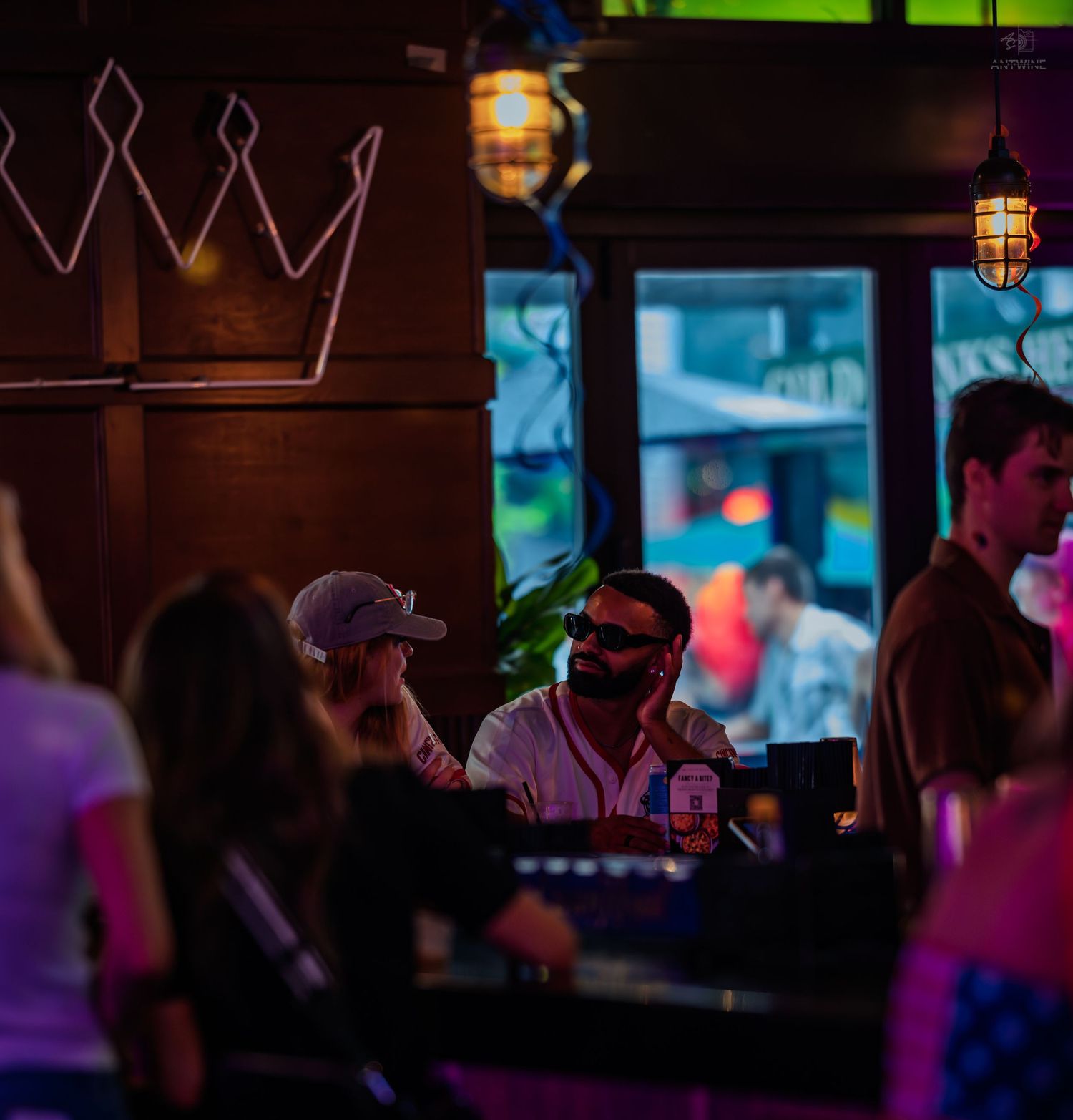 A man in sunglasses and cap sits at a dark bar beneath a neon crown sign.