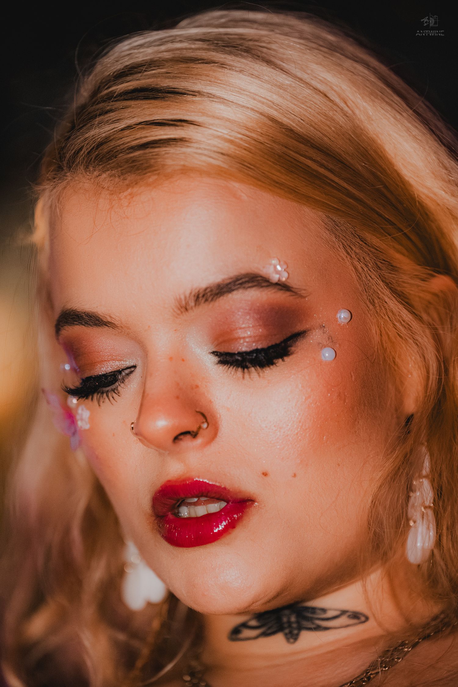 Beautiful blonde woman with glitter face gems, bold red lips, tattoo, and rose gold eye makeup at festival.