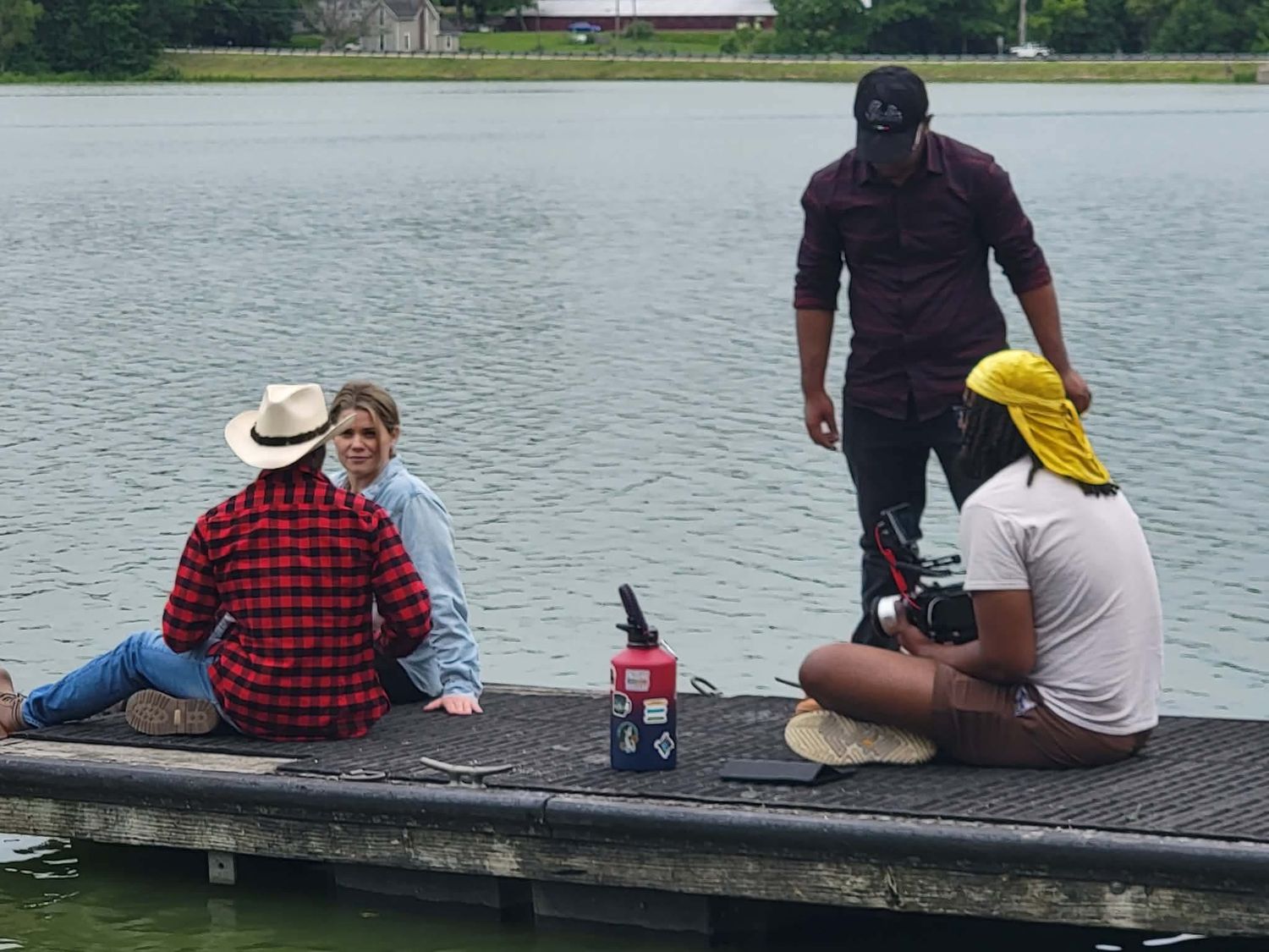 Small film crew sits and stands on a lakeside dock during an outdoor video production shoot.