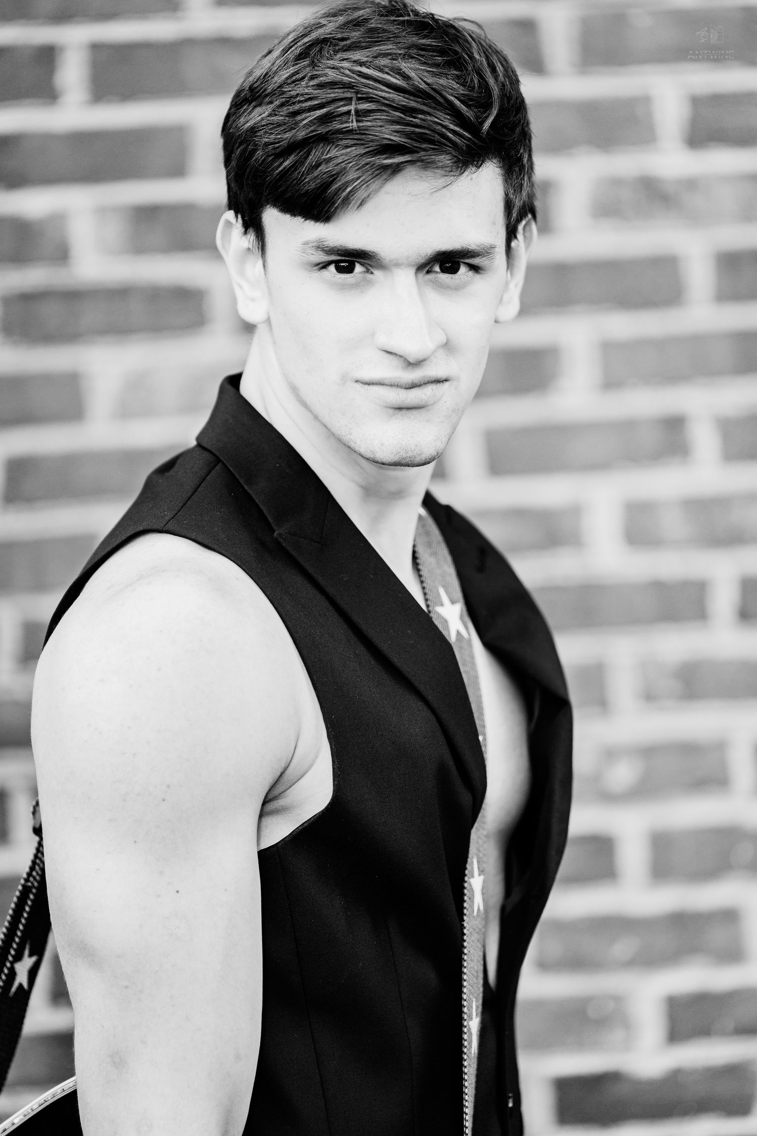 Young man in sleeveless black vest poses against brick wall in black and white photo.