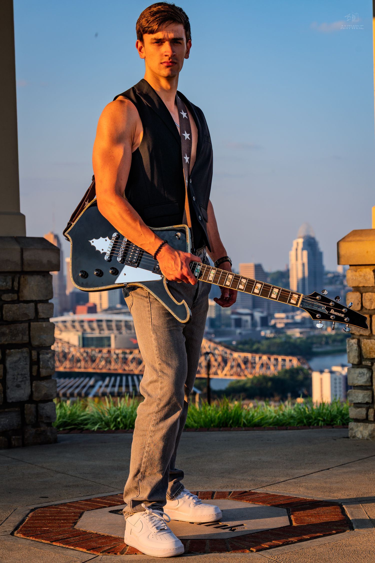 Young male guitarist poses with black electric guitar against a city skyline at golden hour.