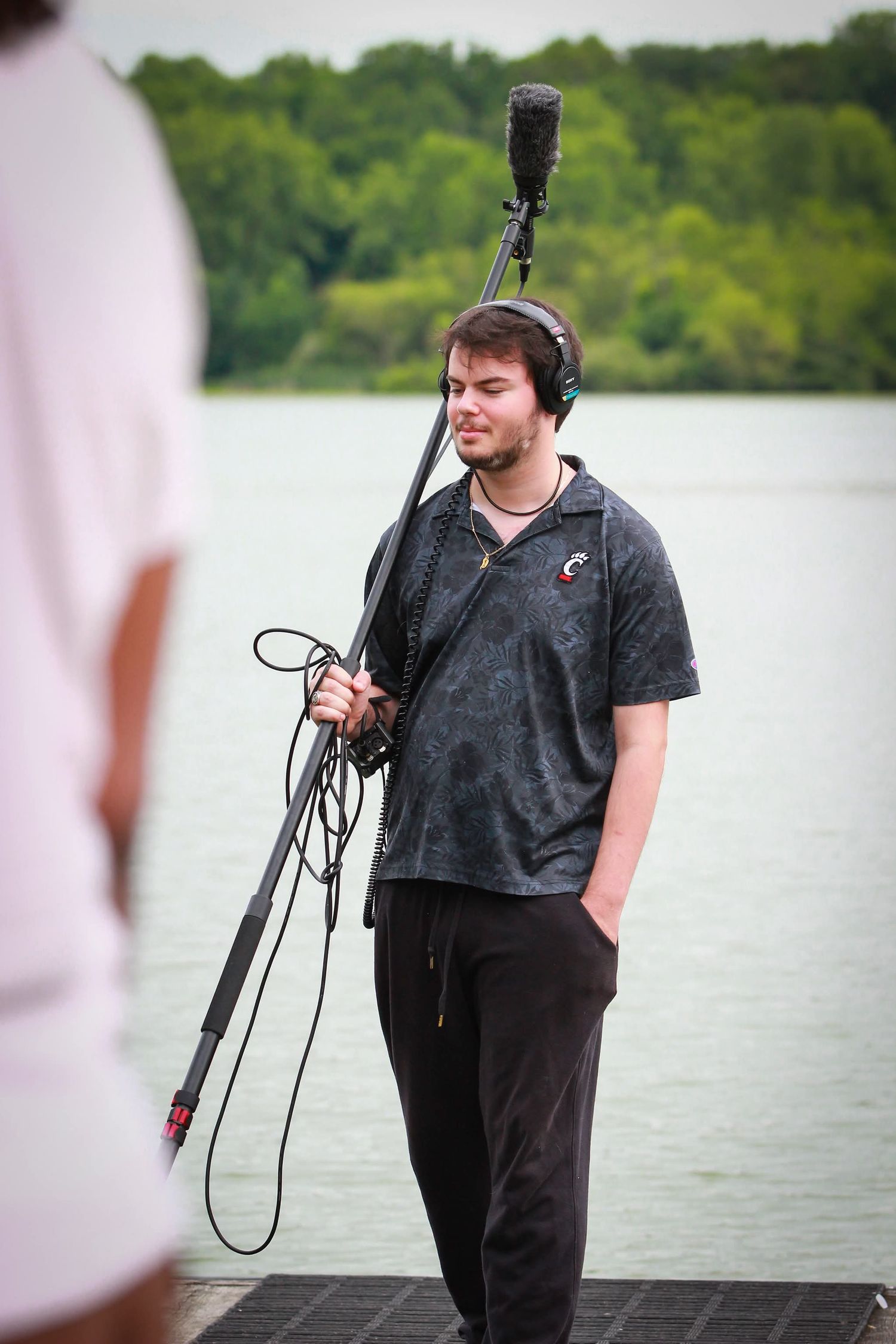 Sound technician holding boom microphone with headphones on outdoor film set.