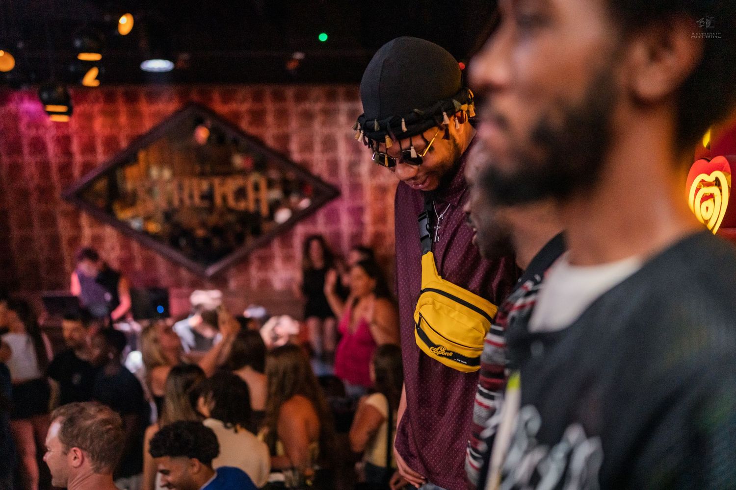 A man in a cap and yellow fanny pack stands at a crowded nightclub with a neon sign backdrop.