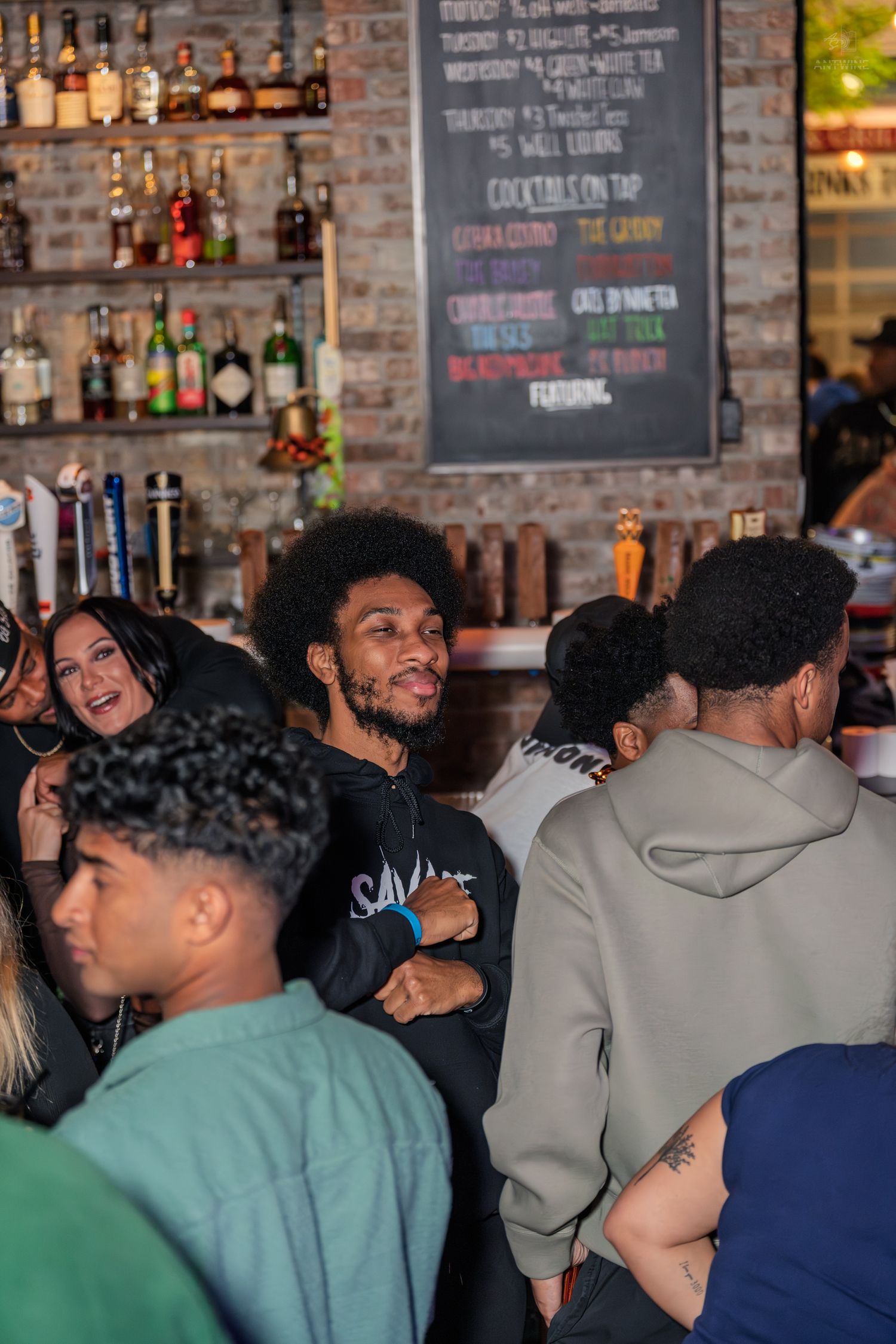 Diverse group of young adults socializing and laughing at a lively bar with cocktail menu on chalkboard wall.