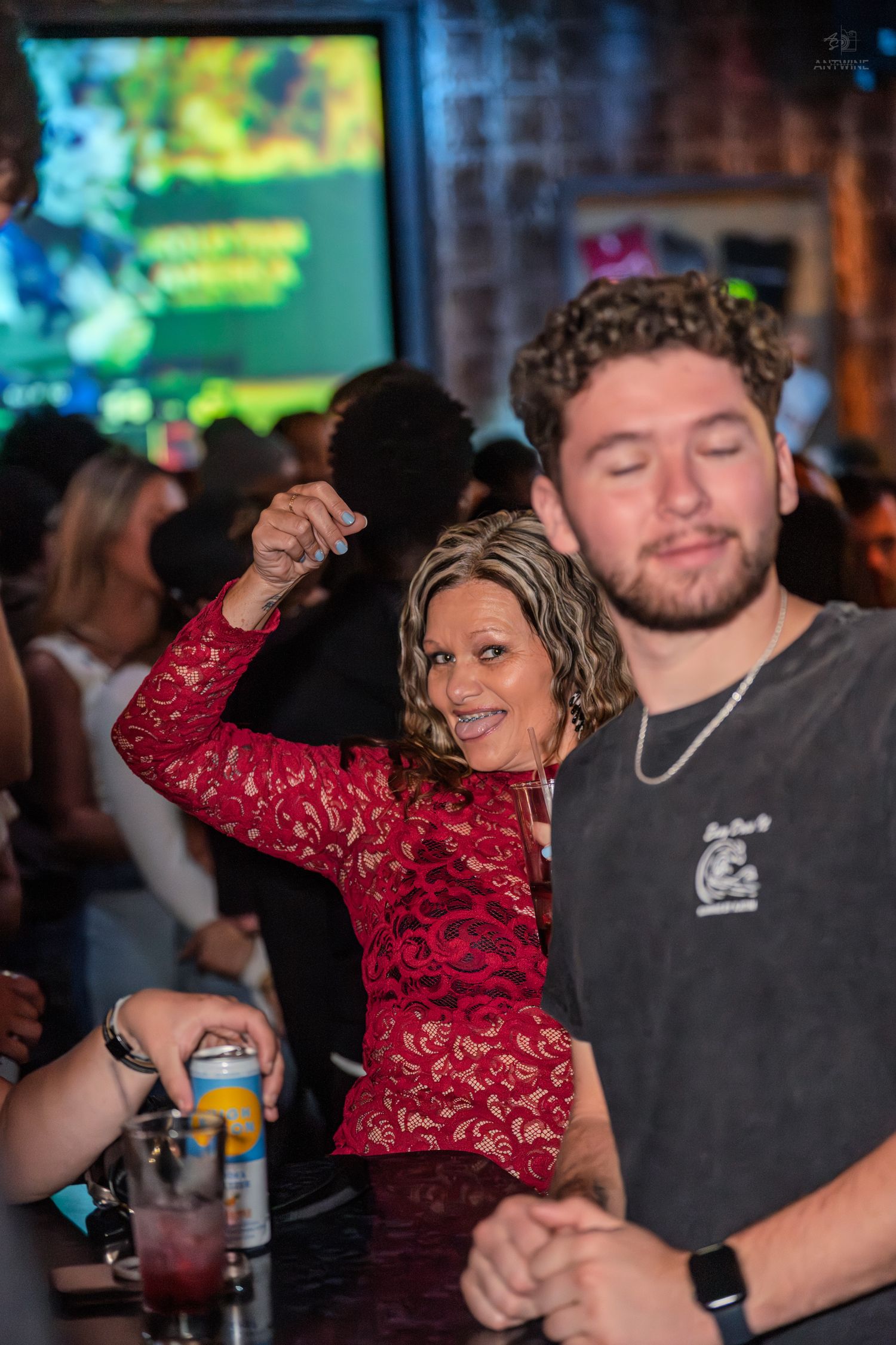 Smiling woman in red lace dress dancing with raised fist next to a man at a nightclub bar.