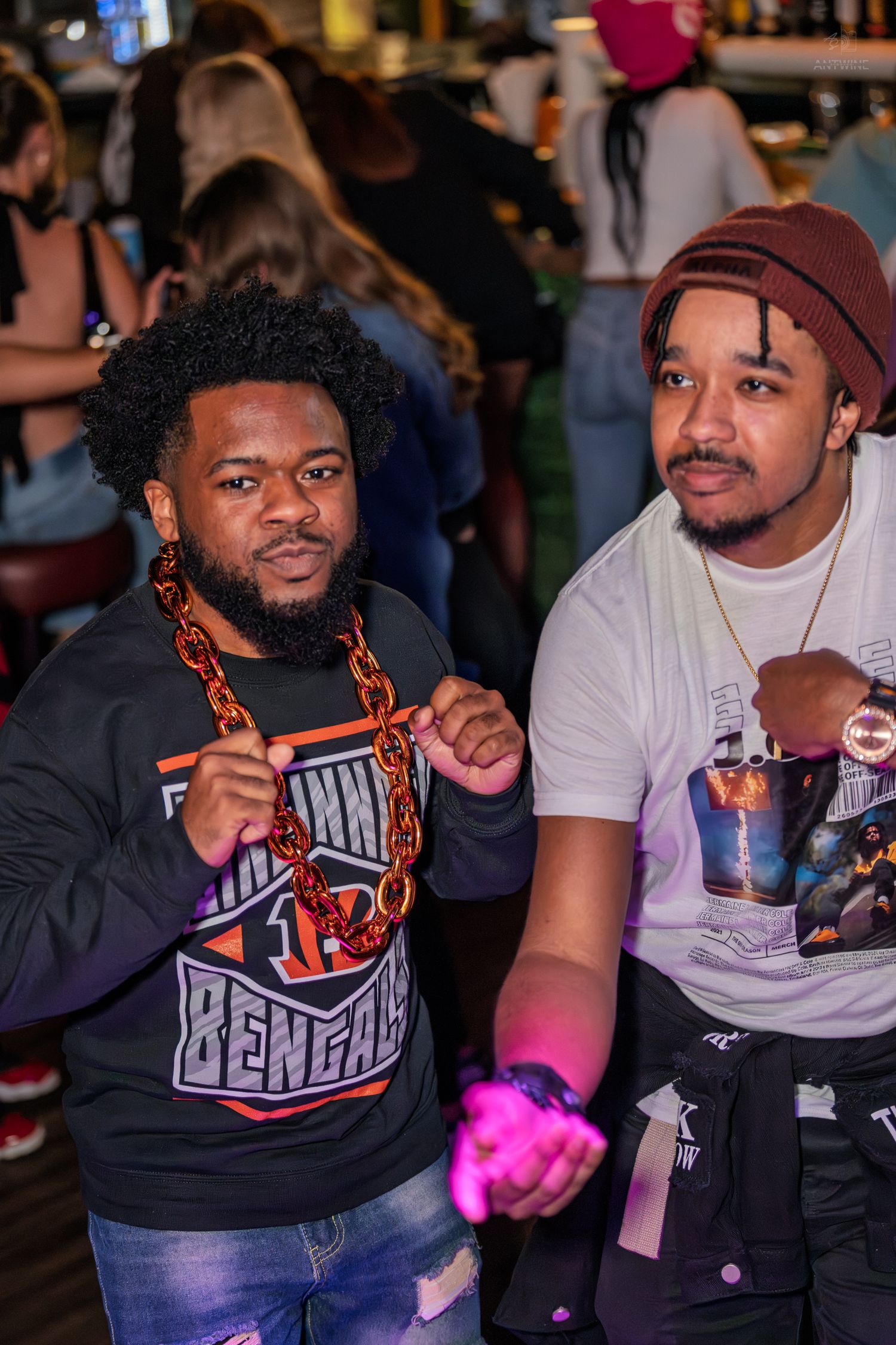 Two men posing with fists up at a bar, one wearing a Bengals jersey and orange chain necklace.