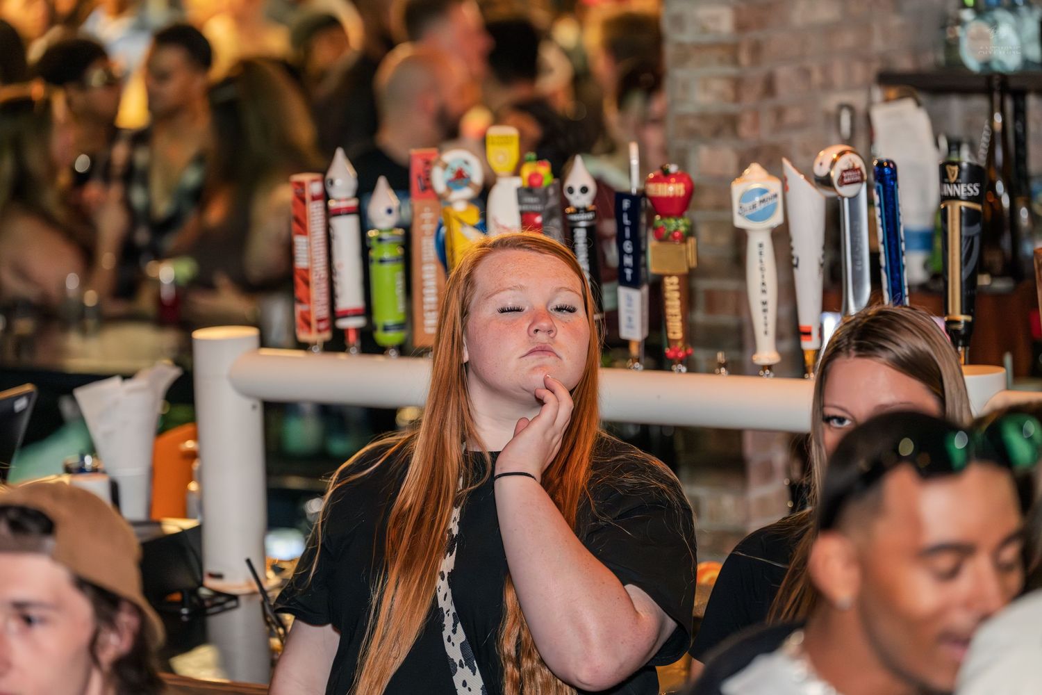 Young redhead woman resting her chin on her hand near a row of beer taps at a bar.
