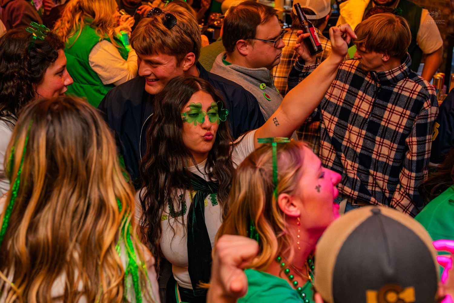 Woman wearing green shamrock sunglasses posing with friends at a festive St. Patrick's Day bar party.