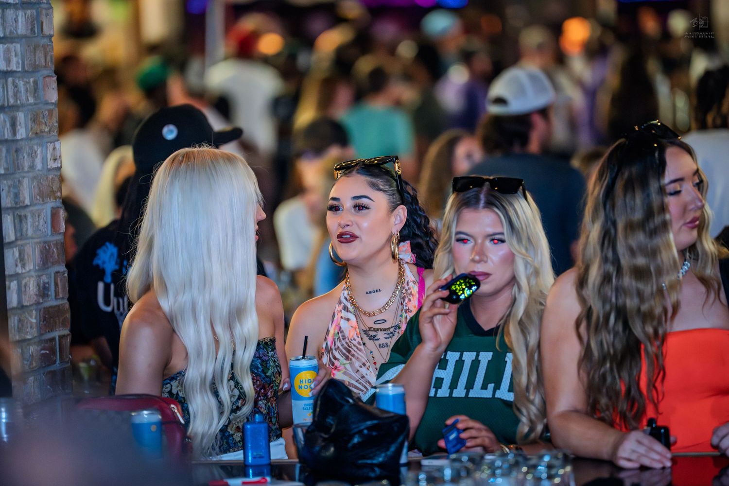 Young women socializing at a crowded nightclub bar, with colorful drinks and vibrant nightlife atmosphere.