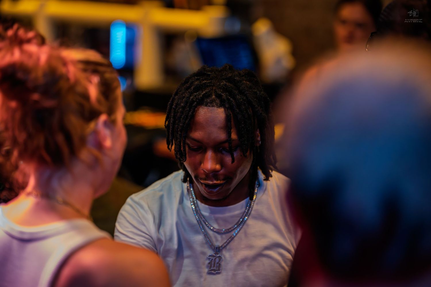 Young man with dreadlocks and chain necklace at a crowded indoor event.