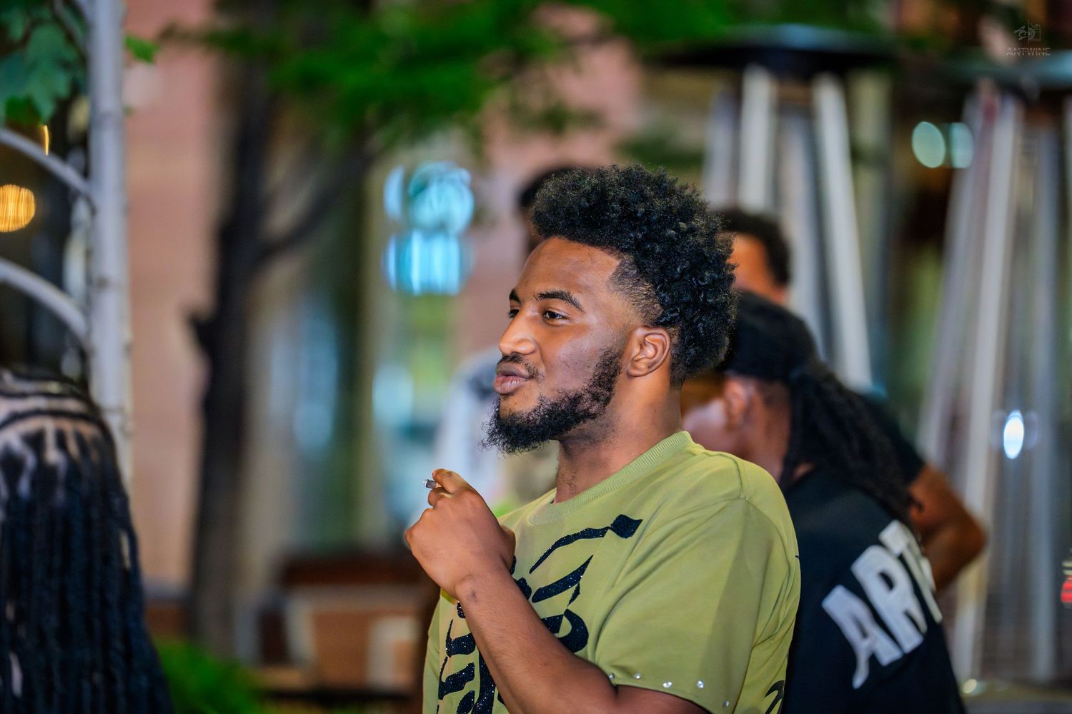 Young man in olive green graphic tee gesturing outdoors at night event.
