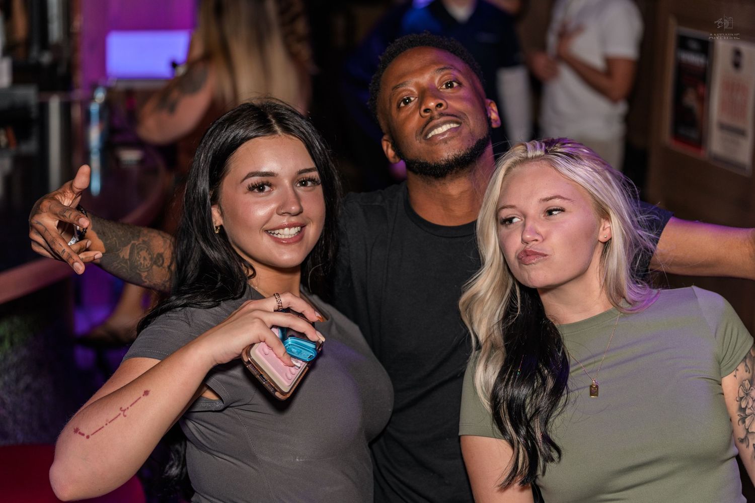 Two women and a man smiling and posing together at a vibrant nightclub or bar event.