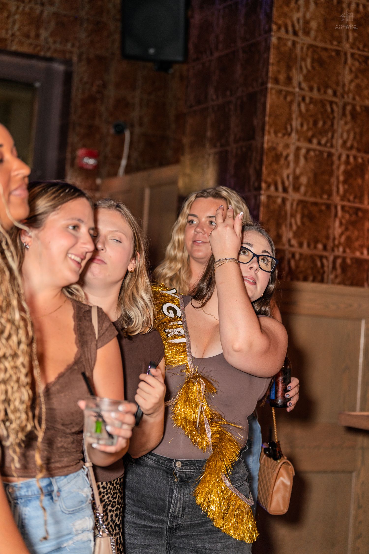 Four young women in a bar, one wearing a gold sash, smiling and watching something off-camera.