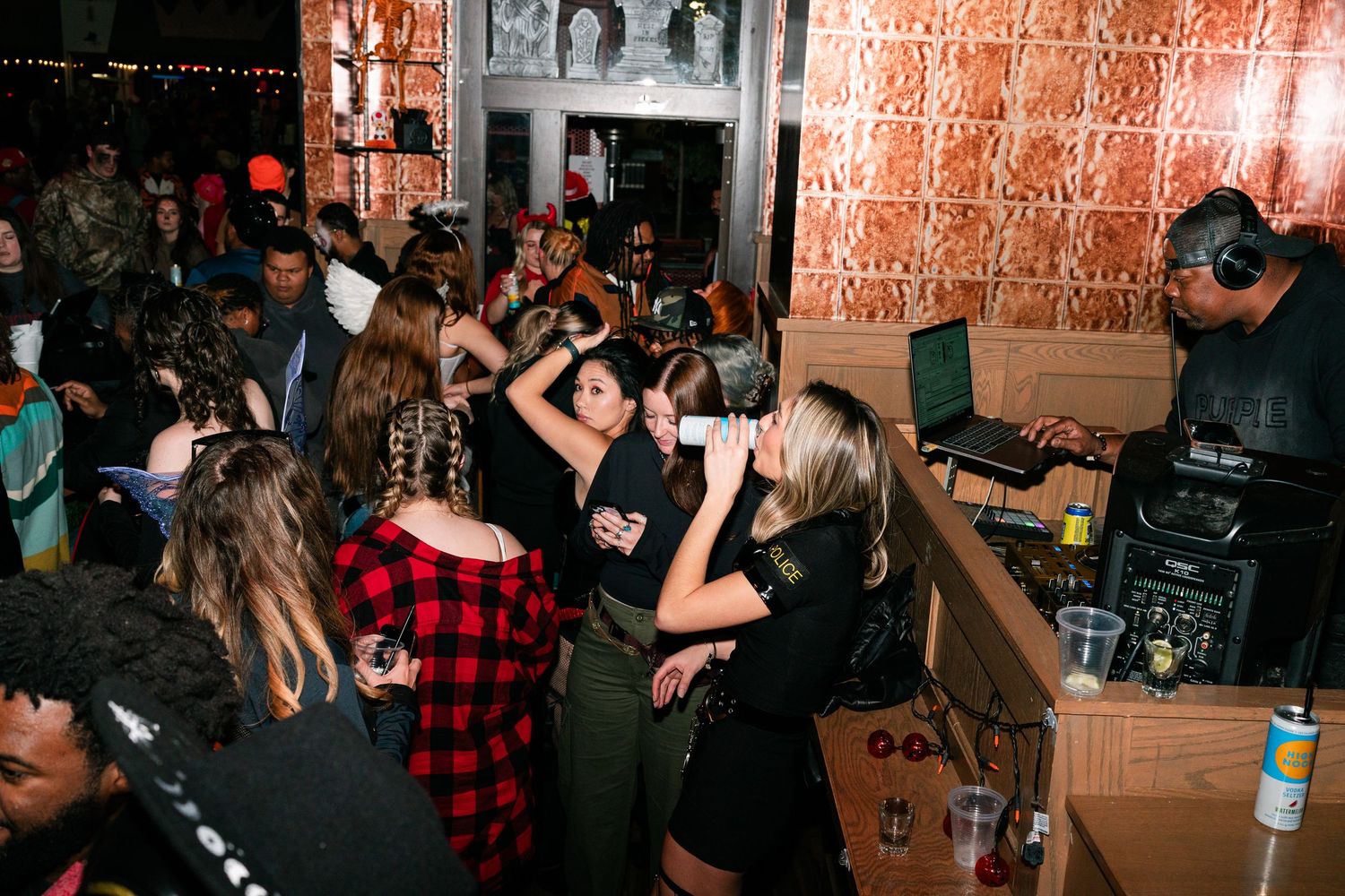 Busy bar scene with guests in Halloween costumes dancing near a DJ booth.