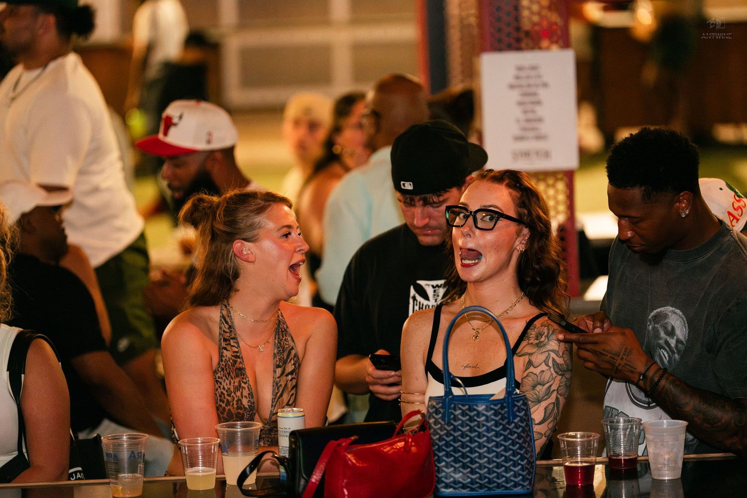 Two tattooed women laugh together at a lively outdoor bar event at night.