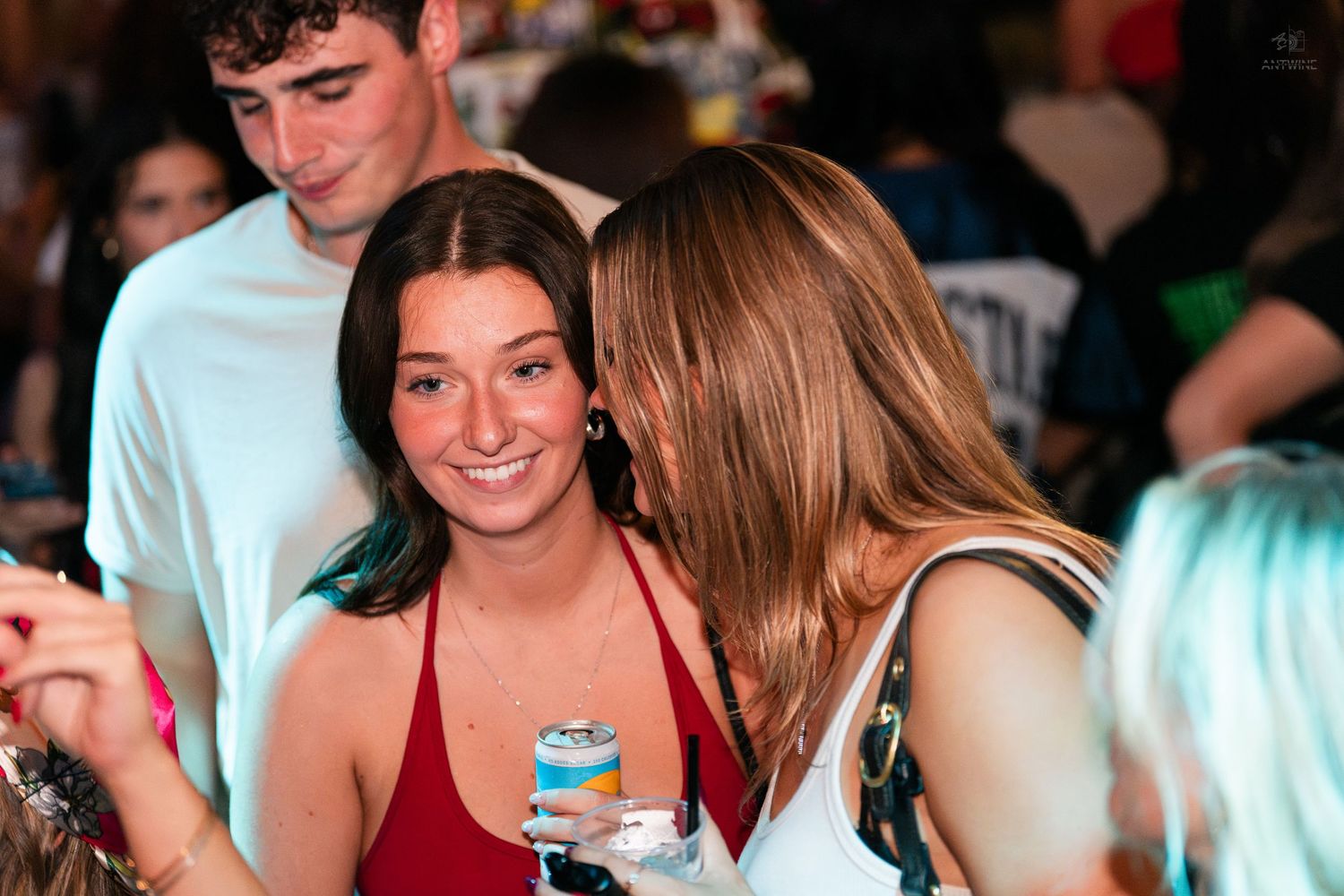 Smiling brunette woman in red top holding a drink can at a crowded party venue.