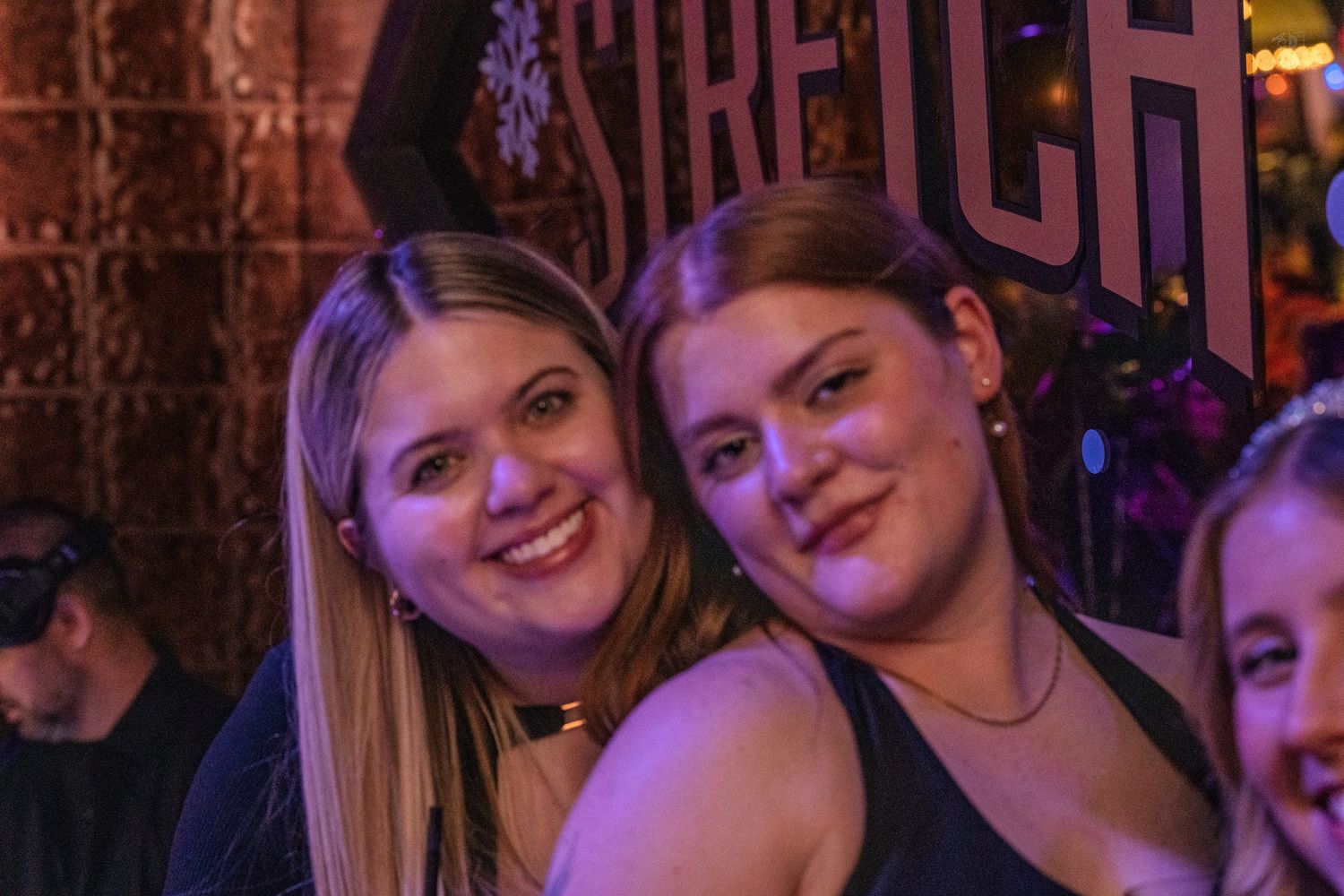 Two smiling women posing together in front of a Stretch bar sign.