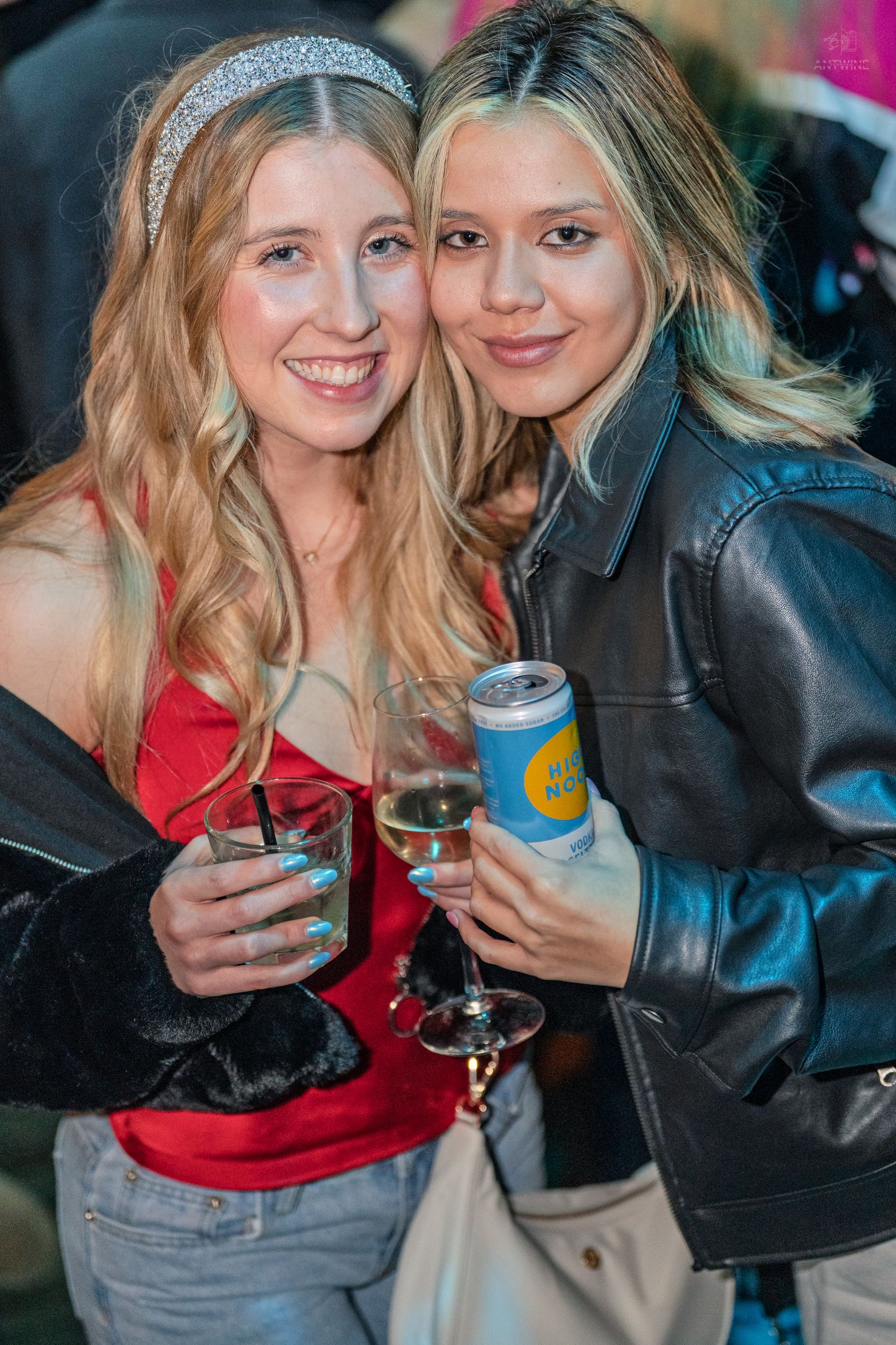 Two blonde women smile holding drinks including a canned beverage at an outdoor night event.