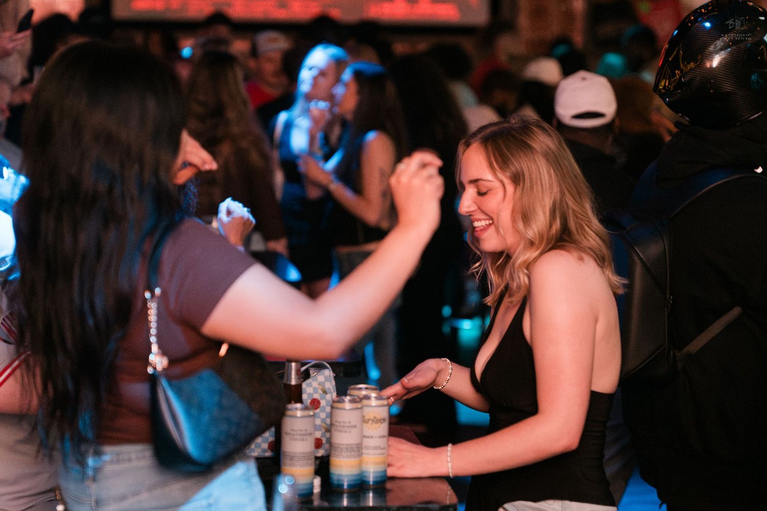 Two women laugh and interact at a bar counter with drinks lined up in a lively nightclub.