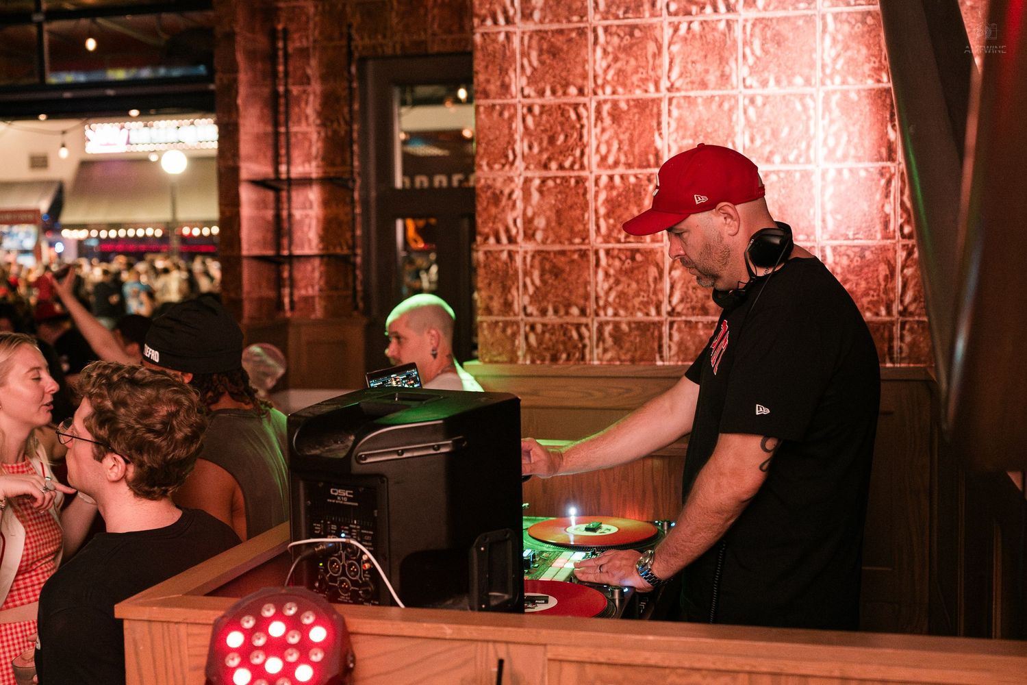 DJ in red cap mixing music at turntables in a lively bar venue.