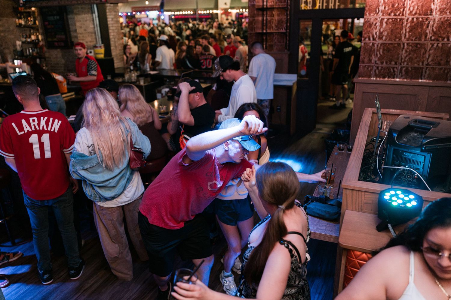 Energetic crowd dancing and socializing inside a lively bar with a DJ setup and neon lights.