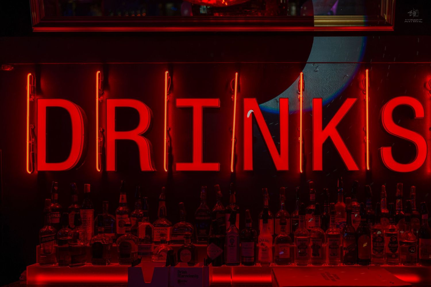 Glowing red neon DRINKS sign illuminating a bar shelf filled with liquor bottles in a dimly lit setting.