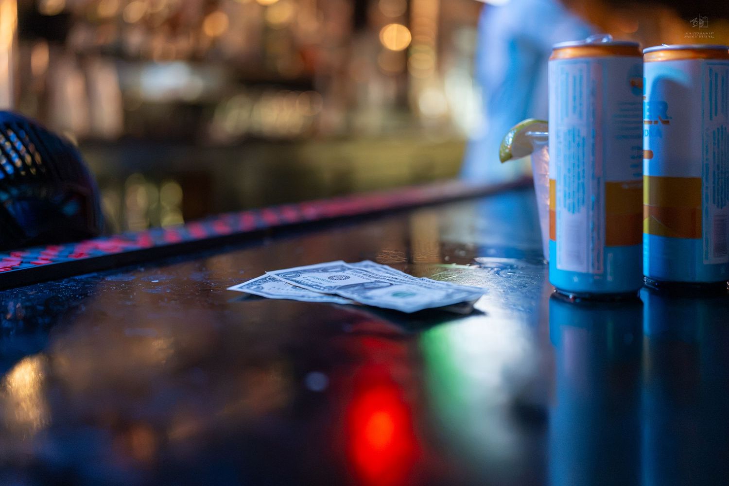 Cash and wallet on a dark bar counter beside two blue energy drink cans in a dimly lit bar.